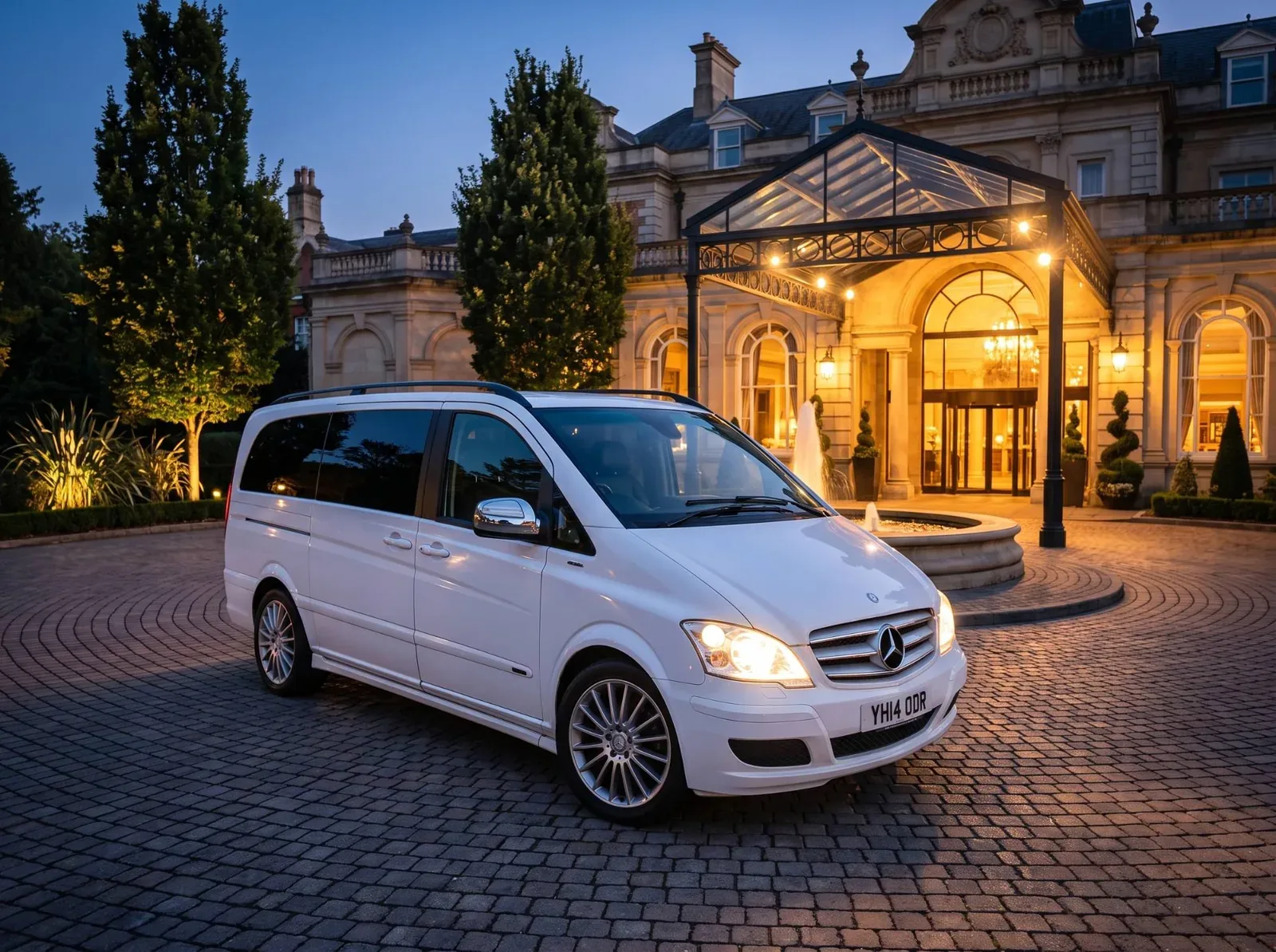 White Mercedes 9-seater at an illuminated hotel entrance at dusk
